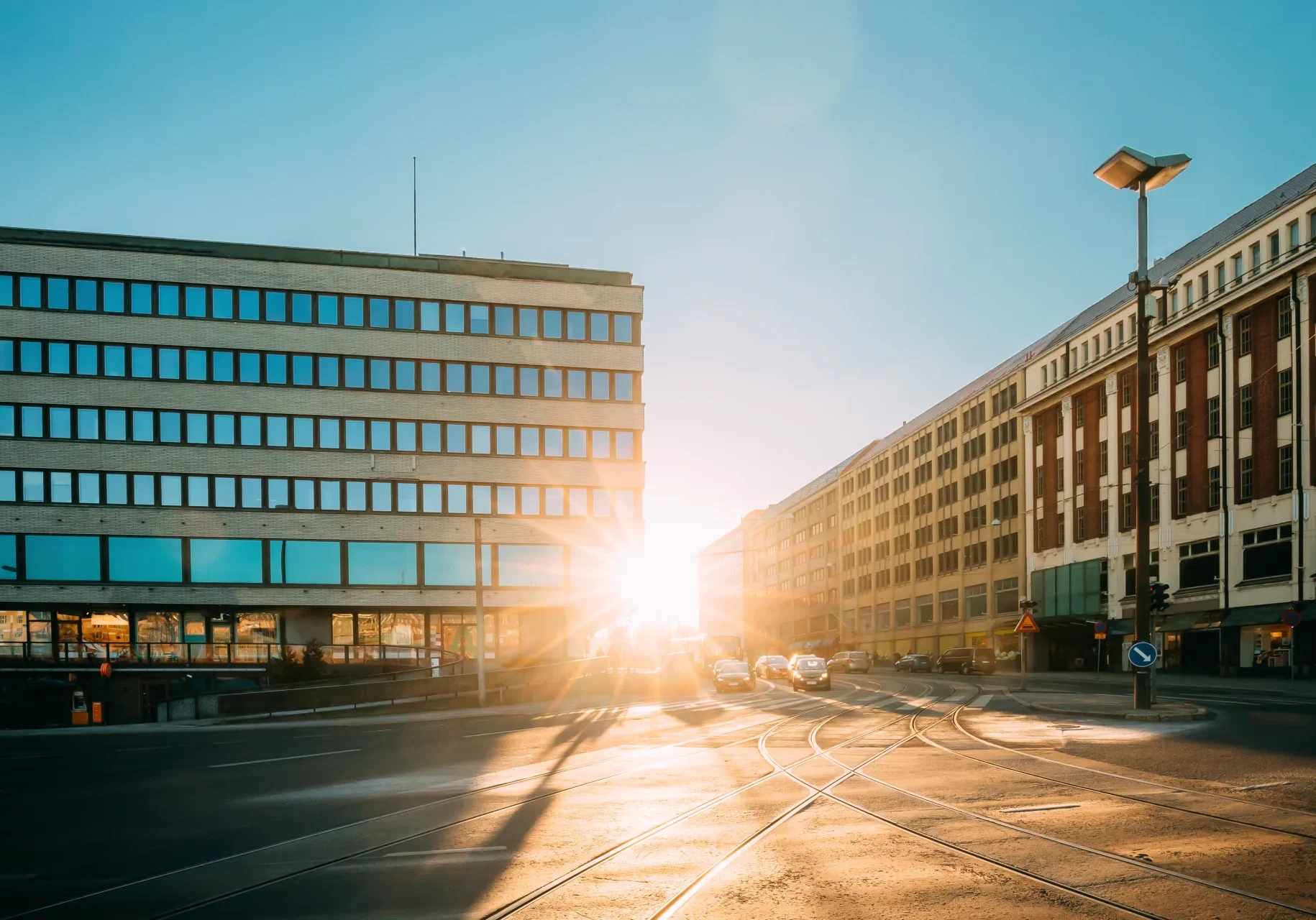 Sollyset skinner sterkt mellom to store kontorbygg og lyser opp en stille industrigate med noen få biler og synlige trikkeskinner, alt under en klar blå himmel.