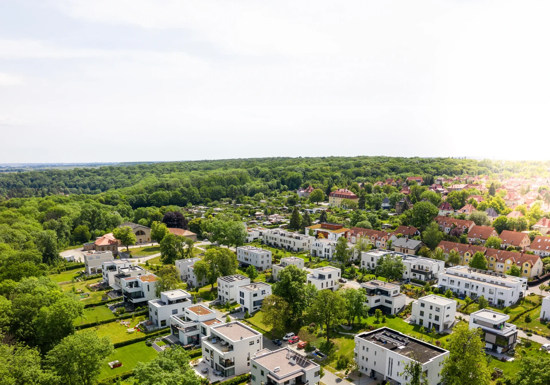 Luftfoto av et forstadsområde med moderne hvite hus omgitt av frodige grøntområder. Scenen inkluderer trekantede gater og en fjern horisont under en delvis overskyet himmel.