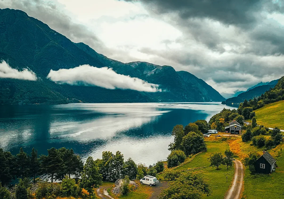 Vakker utsikt over en Serwent-fjord med stille vann som reflekterer den overskyede himmelen. Fjell reiser seg på hver side, utsmykket med grøntområder og skyer som omslutter bakkene. Et lite hus og bobil ligger blant trærne på høyre side, og inviterer til rolig ettertanke.
