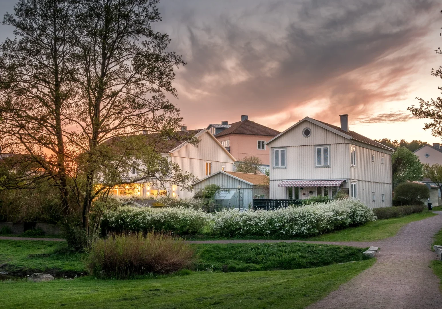 Et fredelig boligstrøk ved solnedgang, med hvite og beige hus omgitt av grønne plener, trær og blomstrende busker langs en svingete sti under en dramatisk himmel.
